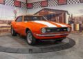 An orange 1968 Chevrolet Camaro SS with white racing stripes, a standout among classic muscle cars, is displayed indoors on a rotating platform against a desert-themed backdrop.