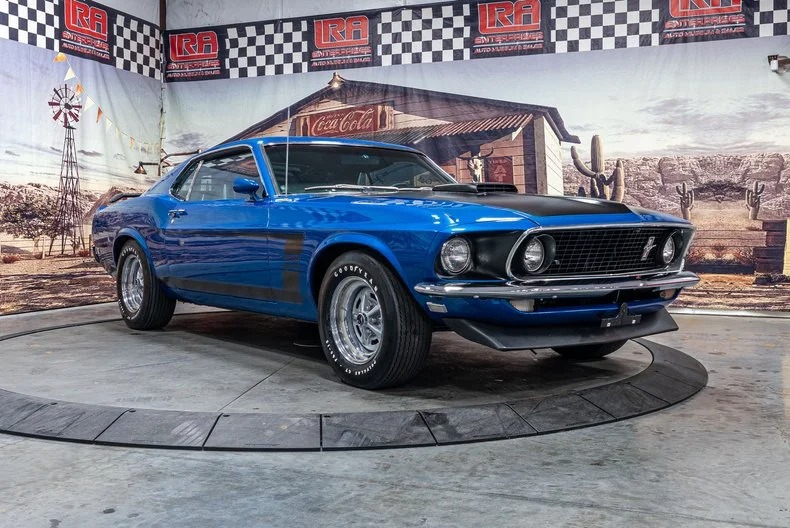 A blue 1969 Ford Mustang Boss 302 coupe, a true icon among Classic Muscle Cars, is displayed indoors on a rotating platform with a mural of a rural scene in the background.