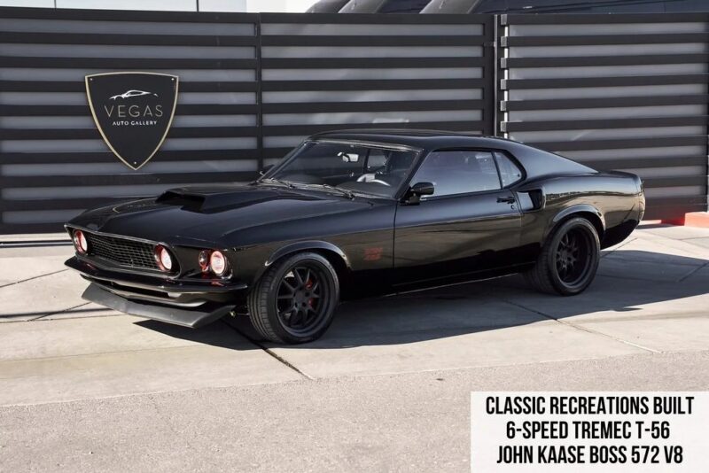A black 1969-style Ford Mustang parked outdoors, featuring custom wheels; text in the corner lists specs including a 6-speed Tremec T-56 and John Kaase Boss 572 V8 engine—a true icon among classic muscle cars.