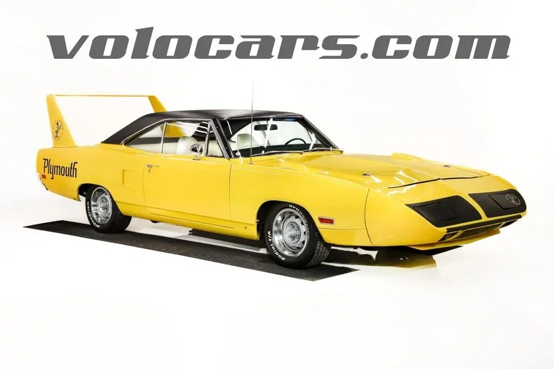 A yellow Plymouth Superbird classic muscle car with a black roof and rear wing is displayed in a studio with "volocars.com" written above.