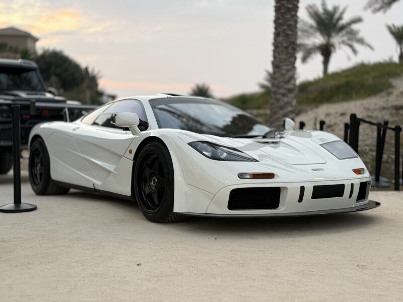 A white McLaren F1 sports car with black wheels is displayed outdoors, surrounded by black stanchions, with palm trees and a sunset sky in the background—an iconic sight reminiscent of the Halo Cars Group collection.