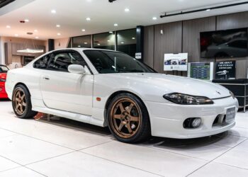 A white Nissan Silvia S15 coupe with bronze wheels is displayed indoors under bright lighting in a modern showroom, offering a chance to own this luxury car.