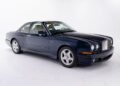A dark blue two-door Bentley luxury car coupe is parked in a white studio, with its beige interior visible through the windows.
