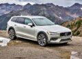 A silver Volvo V60 Cross Country wagon is parked on a gravel area near a mountain range on a cloudy day.