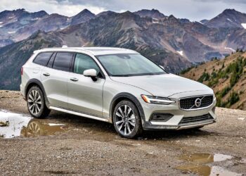 A silver Volvo V60 Cross Country wagon is parked on a gravel area near a mountain range on a cloudy day.
