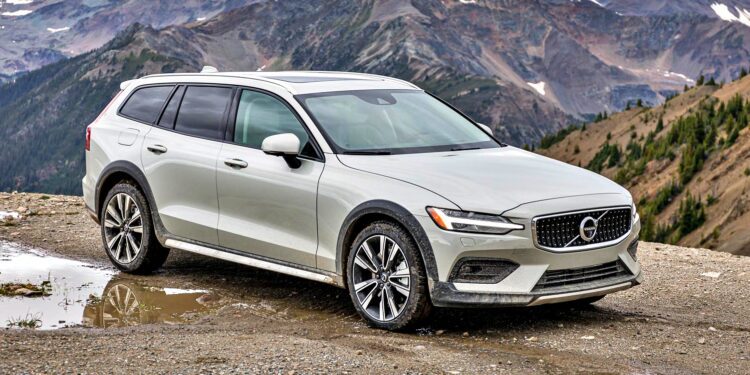 A silver Volvo V60 Cross Country wagon is parked on a gravel area near a mountain range on a cloudy day.