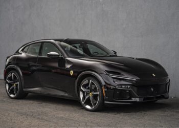 A black Ferrari Purosangue SUV is parked next to a gray concrete wall on a paved surface, viewed from the front left side.