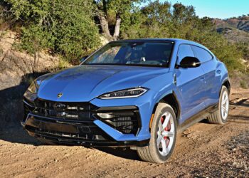 A blue 2025 Lamborghini Urus SE, the heavy hitter delivering plug-in hybrid punch, is parked on a dirt road with red brake calipers, framed by trees and hills in the background.