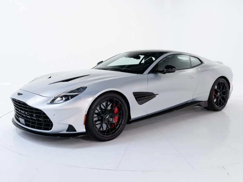 A silver Aston Martin V12 coupe with black wheels and red brake calipers is parked in a white studio setting, viewed from a front-side angle.