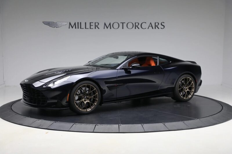A dark blue Aston Martin Vanquish coupe with a powerful V12 engine is displayed in a showroom, highlighted by the "Miller Motorcars" signage in the background.