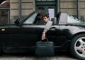 A man sits in a black Porsche Design convertible sports car with the door open, holding a black briefcase, parked in front of a building—a scene worthy of any 2025 Holiday Gift Guide.