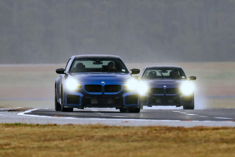 Two blue BMW cars with headlights on drive on a wet race track, the lead pulling ahead. First Drive: 2026 BMW M2 CS Is The Lightest M Car You Can Buy New—showcasing speed and agility in action.