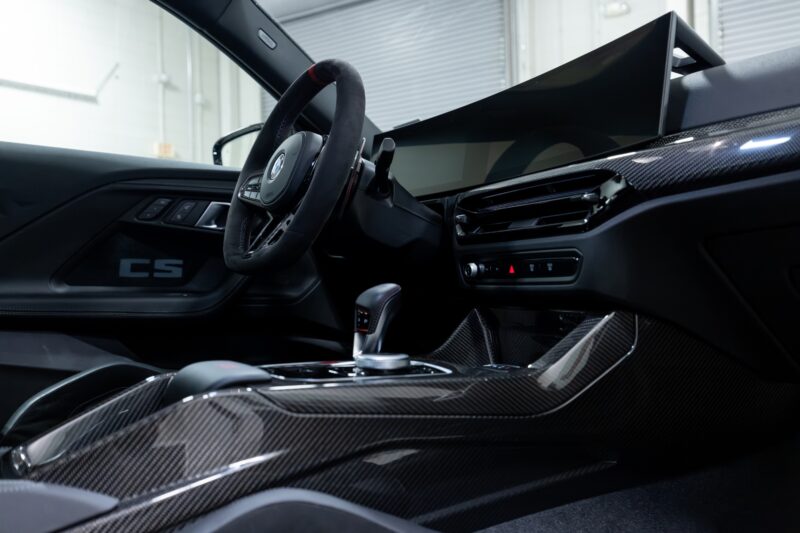 Interior of a modern sports car featuring a digital dashboard, carbon fiber trim, paddle shifters, and a CS badge on the door panel—just like the First Drive: 2026 BMW M2 CS, the lightest new M Car you can buy.