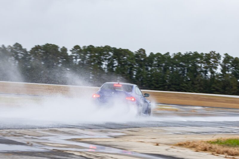 A car drives on a wet track, kicking up water and creating a spray behind it under overcast skies—much like the First Drive: 2026 BMW M2 CS, the lightest M Car you can buy new.