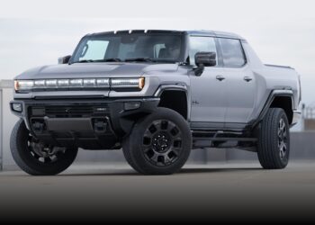 A silver GMC Hummer EV pickup truck, featured in the Review: 2026 GMC Hummer EV Pickup Opens A Toybox Of Excess, is parked outdoors on a concrete surface, viewed from the front left angle.