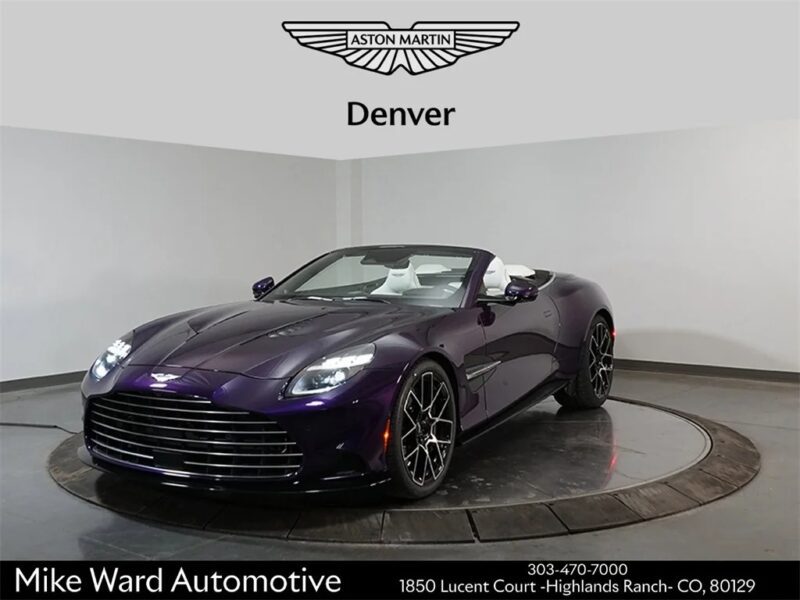 A purple Aston Martin Vanquish convertible is displayed in a showroom with a rotating platform, under signage for Aston Martin Denver and Mike Ward Automotive.