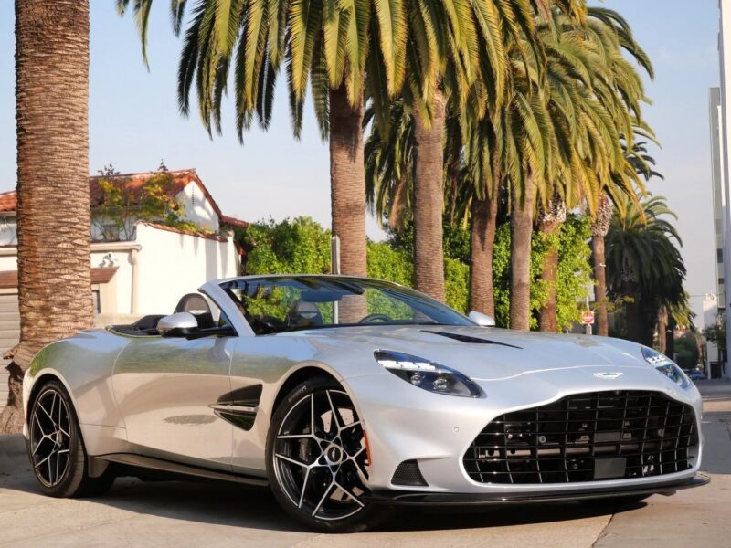 A silver Aston Martin Vanquish convertible sports car parked on a street lined with palm trees, with buildings visible in the background.