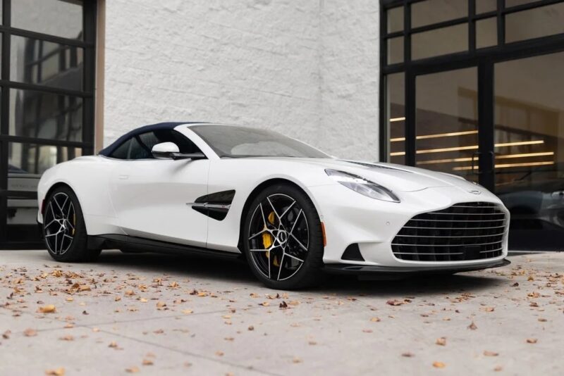 A white Aston Martin Vanquish convertible with black and silver wheels is parked on a concrete driveway near a modern glass building.