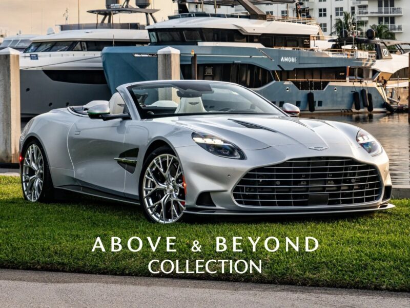 A stunning Aston Martin Vanquish V12 silver convertible is parked on grass near a marina with yachts in the background. Text reads "Above & Beyond Collection.