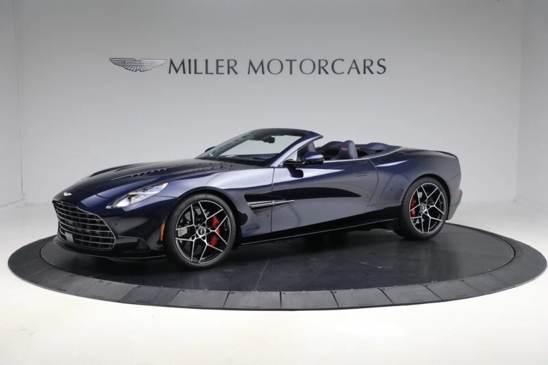 A dark blue Aston Martin Vanquish convertible V12 sports car displayed in a showroom with "Miller Motorcars" signage on the wall behind it.