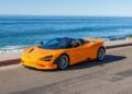 A bright orange luxury car convertible sports car is parked on a coastal road with the ocean and clear sky in the background.
