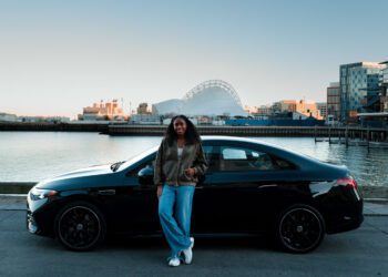 A woman, possibly a Global Brand Ambassador, stands in front of a parked black Mercedes-Benz sedan by the waterfront, with modern buildings and a distinctive arched structure in the background.