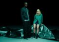 Two people pose in front of a dark background, standing and sitting on a crumpled black tarp. The person on the right sports a green sweatshirt and shorts from the PUMA x Aston Martin F1 Select Collection.
