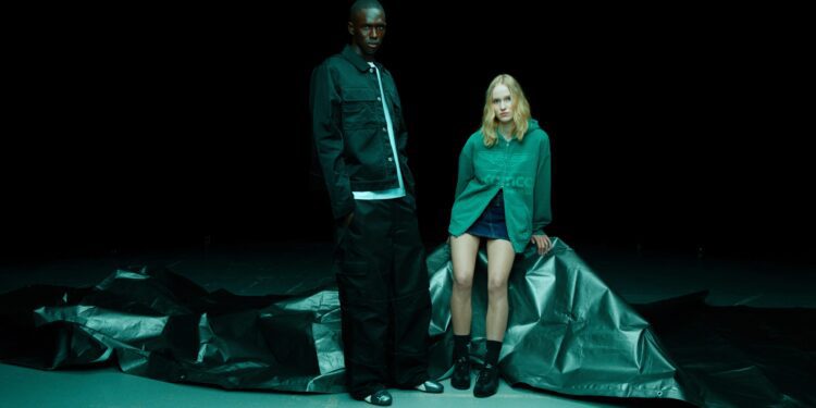 Two people pose in front of a dark background, standing and sitting on a crumpled black tarp. The person on the right sports a green sweatshirt and shorts from the PUMA x Aston Martin F1 Select Collection.