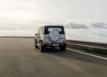 A black Mercedes-Benz SUV with camouflage patterns on the rear drives on a curved highway under a cloudy sky, hinting at the upcoming New G-Class.