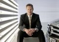 Gorden Wagener, Chief Design Officer of Mercedes-Benz, sits on a metal box beside a Mercedes-Benz car, with vertical fluorescent lights glowing on the wall behind him.