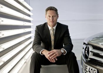 Gorden Wagener, Chief Design Officer of Mercedes-Benz, sits on a metal box beside a Mercedes-Benz car, with vertical fluorescent lights glowing on the wall behind him.