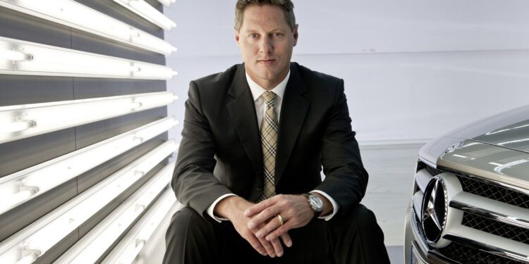 Gorden Wagener, Chief Design Officer of Mercedes-Benz, sits on a metal box beside a Mercedes-Benz car, with vertical fluorescent lights glowing on the wall behind him.