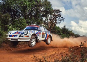 A white Porsche 911 with number 11 on the side speeds down a dirt road in the East African Safari Classic Rally, kicking up dust and lifting off the ground, surrounded by greenery and a cloudy sky.