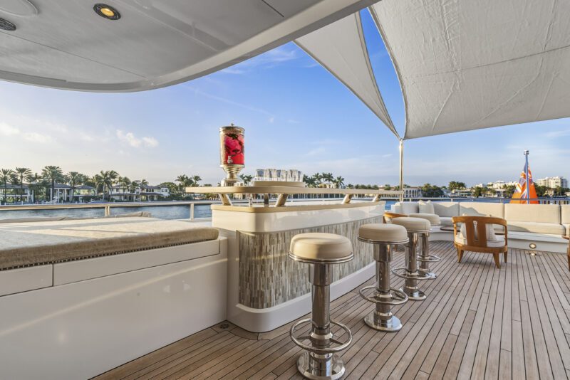 Experience the elegance of a luxury yacht deck aboard Delta Marine Noverra, featuring a shaded bar area with four bar stools, a large glass beverage dispenser, and breathtaking views of palm trees and tranquil water.