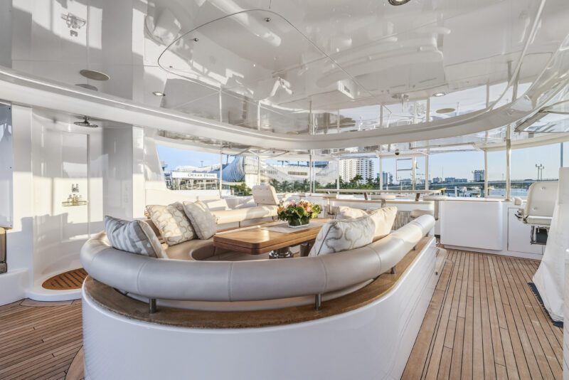 Spacious yacht deck with curved beige seating, a wooden table, decorative pillows, and city buildings in the background—experience five-star living aboard the Delta Marine Noverra luxury yacht charter.