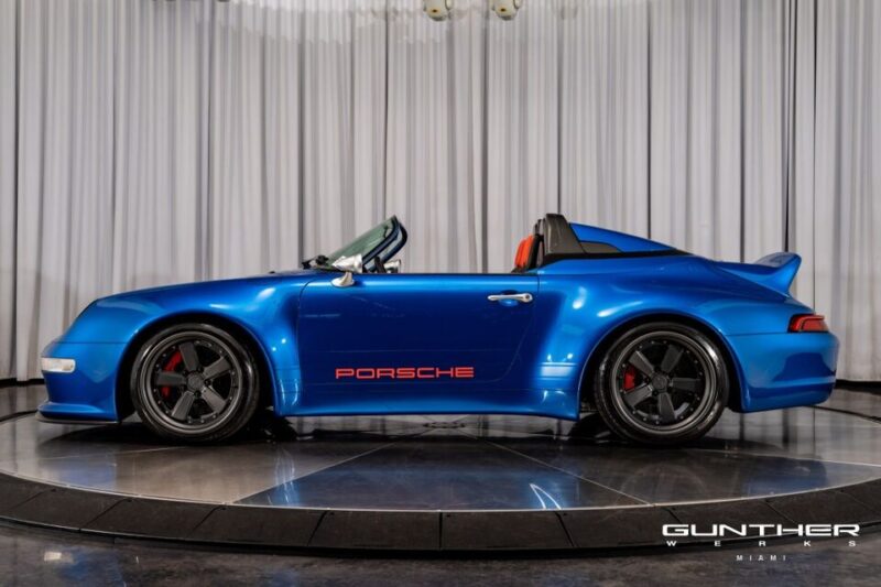 A blue Porsche 911 Speedster convertible with a red interior and black wheels is displayed indoors on a rotating platform, featuring "Porsche" text on the side and Gunther Werks branding in the corner.