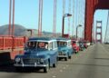 A line of vintage cars and a classic Volvo wagon drives across the Golden Gate Bridge on a clear day, with hills visible in the background.
