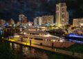 A large, illuminated Delta Marine Noverra yacht docked at a marina at night, with tall city buildings and lights reflected on the water—an iconic scene of five-star living and luxury yacht charter perfection.