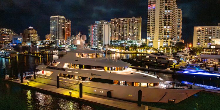 A large, illuminated Delta Marine Noverra yacht docked at a marina at night, with tall city buildings and lights reflected on the water—an iconic scene of five-star living and luxury yacht charter perfection.