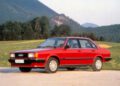 A red classic Audi sedan with the legendary Audi Five-Cylinder Engine is parked on a paved area, set against green fields and forested hills in the background.