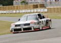 An Audi Quattro race car with its iconic five-cylinder engine makes a turn on a track lined with hay bales, as spectators watch from behind a barrier.