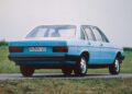 A light blue Audi 100 GL sedan from the late 1970s, featuring an iconic Audi five-cylinder engine, is parked on a paved road beside a grassy field, viewed from the rear left.