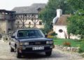 A black Audi 200 sedan with a legendary Audi five-cylinder engine is parked on a cobblestone street in front of historic European buildings and a small chapel, surrounded by trees and flowers.