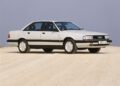 A white Audi sedan from the 1980s, famed for its Audi five-cylinder engine, is parked on a dirt road under a clear sky, viewed from the front left side.