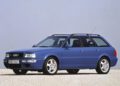 A blue Audi RS2 Avant station wagon, celebrating the legacy of the iconic Audi five-cylinder engine, is parked on a gravel surface against a plain light background.