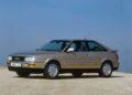 A beige 1990s Audi coupe, featuring an iconic Audi five-cylinder engine, is parked on a flat dirt surface with a clear blue sky in the background.