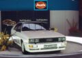 A white Audi Quattro coupe featuring the iconic Audi five-cylinder engine is displayed on a platform with flowers nearby, under an Audi sign at an auto show celebrating the 50th anniversary.