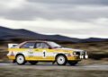 A yellow and white rally car with the number 1 on the door, powered by a legendary Audi five-cylinder engine, drives quickly on a paved road, with blurred mountains in the background.