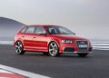 A red Audi RS3 hatchback with its iconic Audi five-cylinder engine is parked on a racetrack, mountains rising in the background—celebrating the 50th anniversary of this legendary Audi engine.