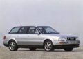 A silver Audi 100 Avant station wagon with the iconic Audi five-cylinder engine is parked on a gravel surface, shown from the side against a plain, foggy background.